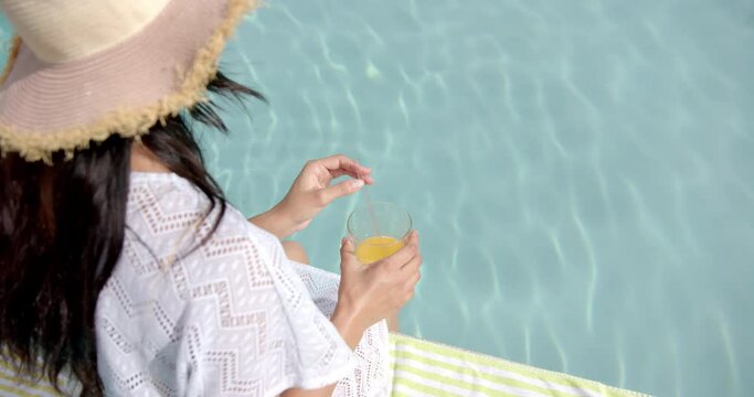 Biracial teenage girl in sunhat drinking juice by pool, copy space, slow motion
