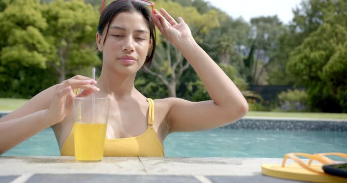 Portrait of happy biracial teenage girl standing in sunny swimming pool with juice, slow motion