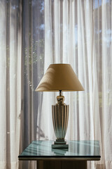 Elegant white antique table lamp with fabric lampshade on the glass table near the window. Lamp silhouette on the curtain in sunny day. Classic decoration for home or hotel architecture