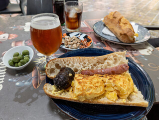Beautiful image of a typical Valencian lunch called esmorzar, a potato omelette sandwich with sausage, blood sausage and all i oli with its beer, soft drink, peanuts and olives in Valencia city, Spain