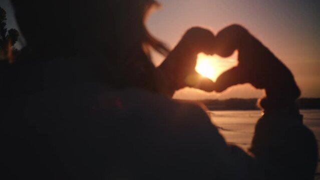 Winter woman silhouette showing heart shape hands in mittens sky bright sun sunset sunrise closeup