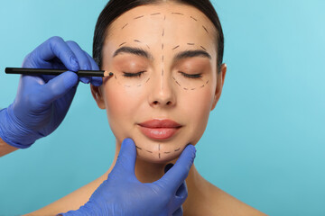 Doctor with pencil preparing patient for cosmetic surgery operation on light blue background,...