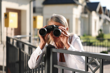 Concept of private life. Curious senior woman with binoculars spying on neighbours over fence...