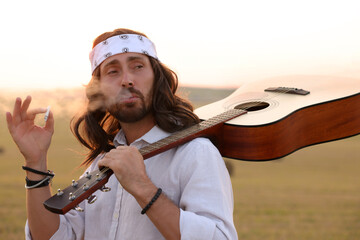 Stylish hippie man with guitar smoking joint in field
