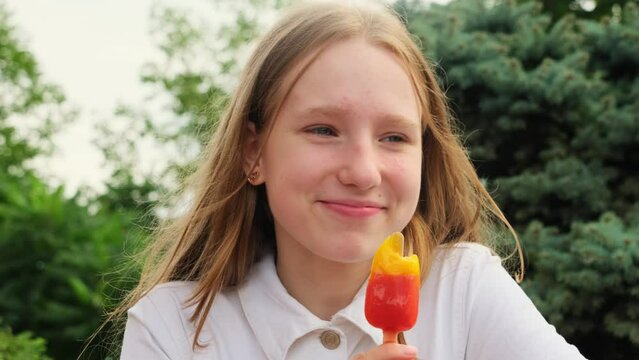 Happy girl eats fruit ice cream in green summer park closeup. Funny teenager bites tasty popsicle resting in city garden slow motion