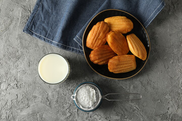 Madeleine cakes in bowl, glass of milk, flour and towel on gray background, top view