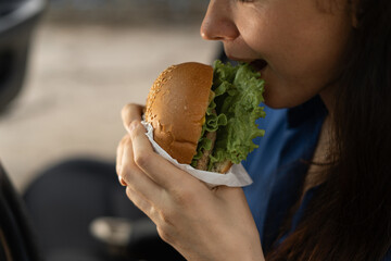 Woman eats burger in car. Bun with cutlet. People travels, having snack on road without leaving car