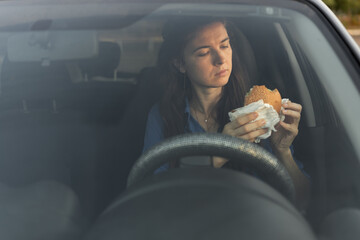 Young hungry woman eats burger while sitting in car, travels, having snack on road. Tourist