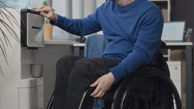 Tilt up shot of young Caucasian wheelchaired man pouring fresh still water from water dispenser into cup while during workday in modern office