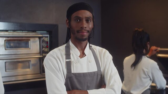 Tilt up portrait of cheerful African American head chef posing for camera while working at restaurant kitchen with colleague