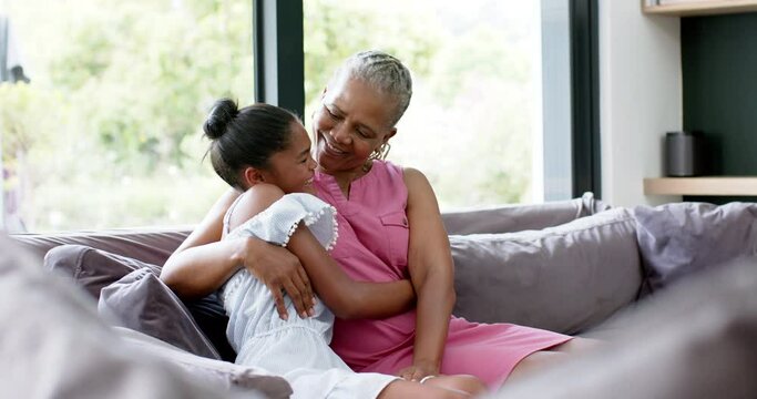 Happy african american granddaughter and grandmother hugging on sofa, copy space, slow motion