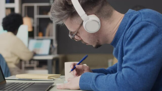 Medium closeup of young Caucasian man in wheelchair sitting by laptop computer in wireless headphones and taking notes in copybook while working with African American female colleague in office