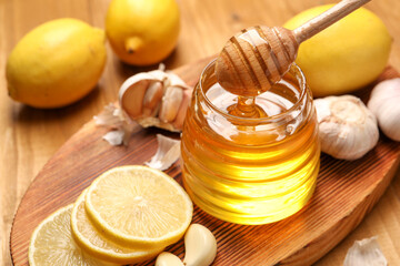 Pouring honey from dipper into jar, garlic and lemons on wooden table, closeup