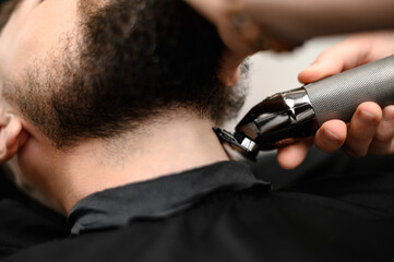 A barber in the salon trims the beard of a man with a trimmer in the neck area
