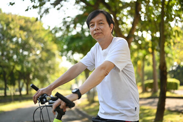 Portrait of happy retired man riding electric bicycle in public park on sunny day. Healthy...