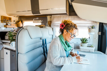One woman sitting inside modern motorhome alternative tiny house and write travel notes on the table. People living vanlife on the road life and vacation. Traveler female indoor leisure van activity - obrazy, fototapety, plakaty
