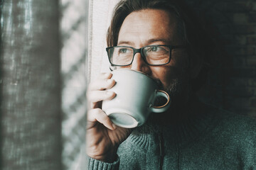 One man with thoughtful and serene expression on face drinking coffee or tea alone at home looking outside the window. Mature male people alone. Healthy mindful lifestyle concept. Wearing glasses - obrazy, fototapety, plakaty