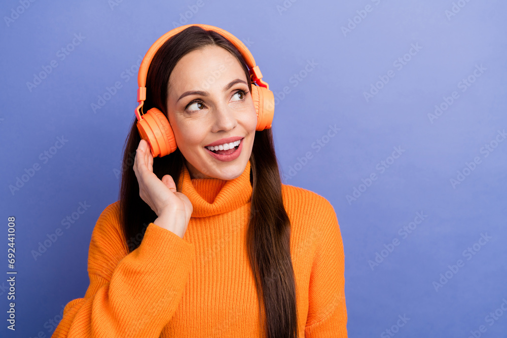 Sticker Photo of charming lady in orange pullover touching headphones look empty space new playlist in spotify isolated on purple color background