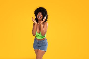 African american happy woman listening to music via wireless headphones with big smile on her face, dancing on yellow studio background.