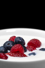 Close-up of a bowl with natural white yogurt and fresh raspberries, blueberries, strawberries on a black background, macro photography. Delicious healthy breakfast. Vertical photo