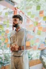 Multiracial colleagues brainstorm business ideas, discussing project costs and planning. Creative and diverse, they share knowledge, writing on sticky notes on a glass wall in a corporate office.