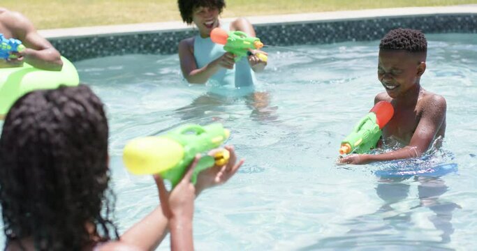 Happy african american parents, son and daughter playing with water pistols in pool, slow motion