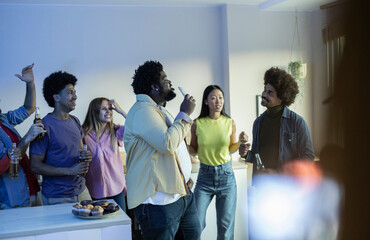 Group of happy multiethnic friends singing karaoke during a party in the apartment, toasting and...
