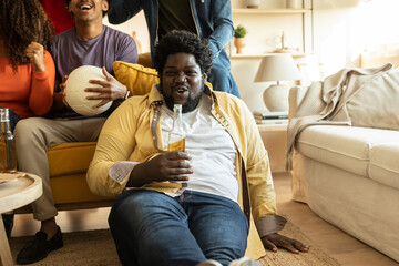 Overweight African American young adult sitting on the floor with his beer in his hand celebrating...