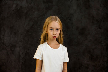Naklejka na ściany i meble Portrait of frowning cover girl kid model 6 year old in white t-shirt expression emotion, angry looking at camera. Scowl child posing at black, studio shot. Kids emotional concept. Copy ad text space