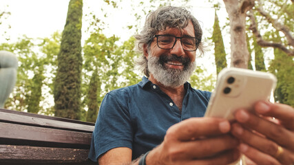 Close-up of smiling elderly man in glasses using phone while sitting on park bench