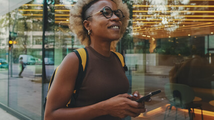Fototapeta na wymiar Smiling young woman in glasses, wearing brown top with backpack on her shoulders, walking down the street with smartphone, texting, chatting with friends on social media