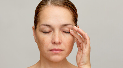 Middle aged caucasian woman of 40s with closed eyes holding finger on eyelid to show eyes aging on...