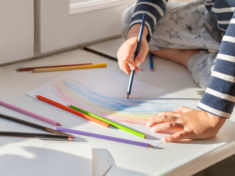 Little boy sits on window sill and draws rainbow with colored pencils. Creative leisure activity with crayons for children at home.