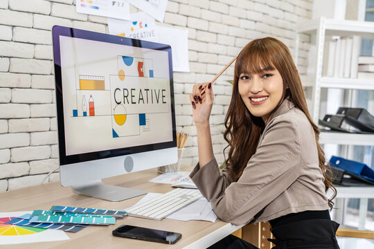 Asian young woman graphic designer smiling and looking at camera sitting at the desk with pc and tablet computer while working in office, Artist Creative Designer Illustrator Graphic Skill Concept