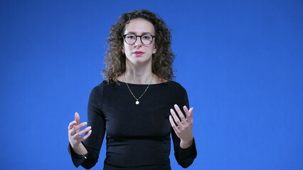 Woman speaking toward camera while standing on blue background. 20s female person looking at camera...