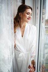 Beautiful young bride in white negligee standing near the window.