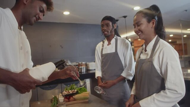 Medium long team of friendly multi-ethnic chefs celebrating end of working day at restaurant and pouring wine into glasses