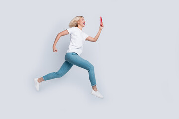 Fototapeta na wymiar Full size photo of cheerful woman dressed white t-shirt denim pants fast run look at smartphone isolated on white color background