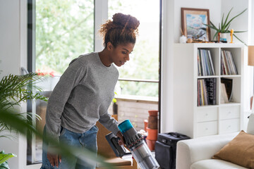 Gorgeous curly concentrated woman is using vacuum cleaner while cleaning her floor - obrazy, fototapety, plakaty
