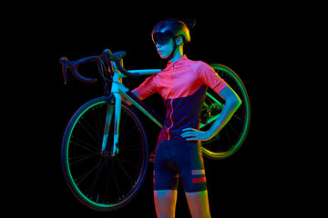 Young man in sportswear, helmet and googles holding bicycle on shoulder isolated over black studio background in neon light - obrazy, fototapety, plakaty