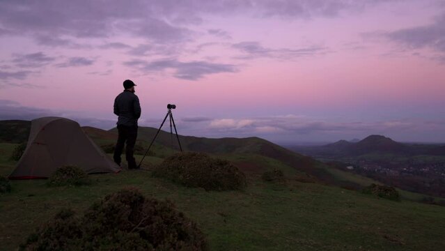A photographer standing beside his camera tripod and wild camping tent in the English hills at dusk