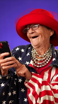 Vertical video of funny crazy happy elderly old toothless woman taking selfie using mobile cell phone wearing US flag jacket and glasses isolated on solid purple background studio portrait. 