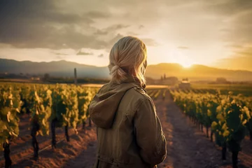Fototapeten woman with blonde hair, wearing a jacket, stands in a vineyard at sunset, with rows of grapevines and mountains in the background, ai generative © larrui