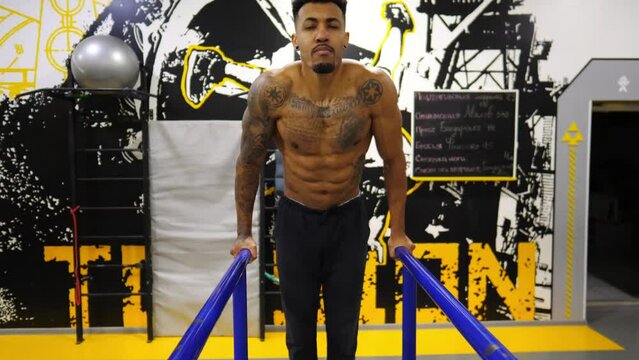 Strong african man doing push ups on parallel bars at sport club. Muscular afro american sportsman with tattoos doing hard exercises modern gym. Concept of healthy and active lifestyle. Slow motion