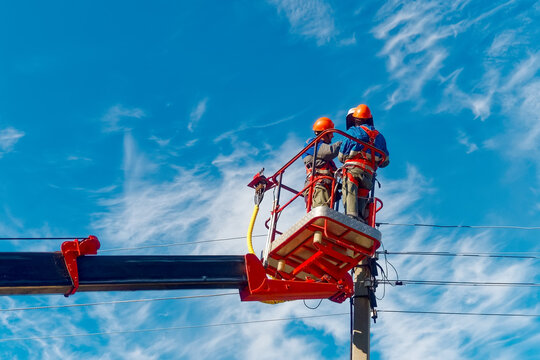 Two electricians from cradle of aerial platform or crane are repairing street lighting lamp. Professional electricians wearing helmets, overalls and insurance work at heights. View of workers from