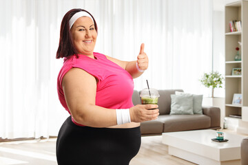 Plus size woman in sportswear holding a green smoothie and gesturing thumbs up