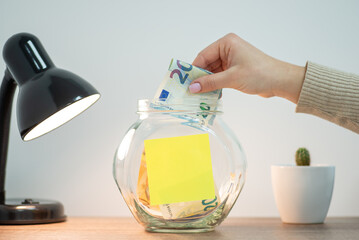 Transparent money box, lamp and small kaktus standing on the table. Female hand putting 20 euro banknote into the glass jar, paper sticker with copy space