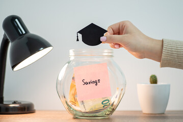 Female hand holding image of black students hat above the transparent money box, close up. Concepts...