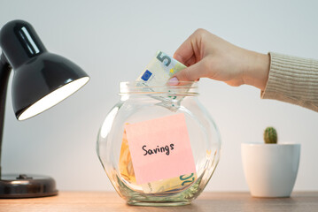 Savings in the glass jar standing on the table. Close up of female hand putting five euro banknote...