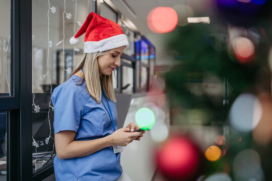 Portrait of beautiful nurse with christmas hat in hospital corridor, holding smartphone. Working in hospital on Christmas day, Christmas Eve. Female doctor working a Christmas shift and can't be with